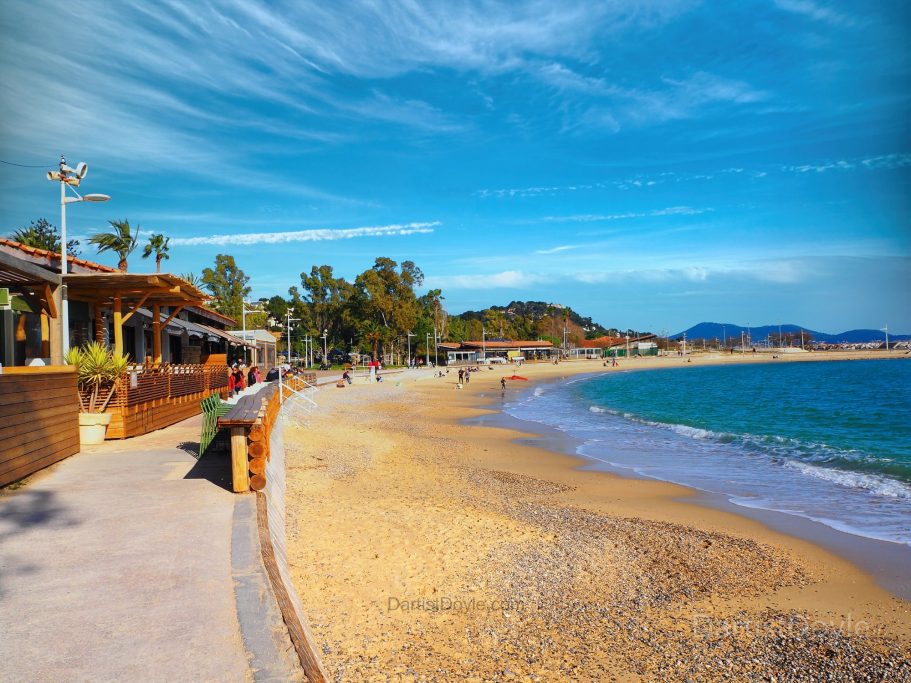 Plage ensoleillée avec sable fin, restaurants en bord de mer et ciel bleu dégagé.