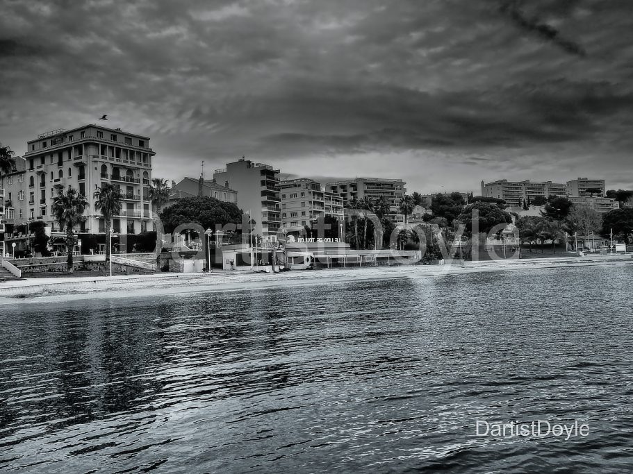 Vue en noir et blanc d'un bord de mer avec des bâtiments et un ciel nuageux.