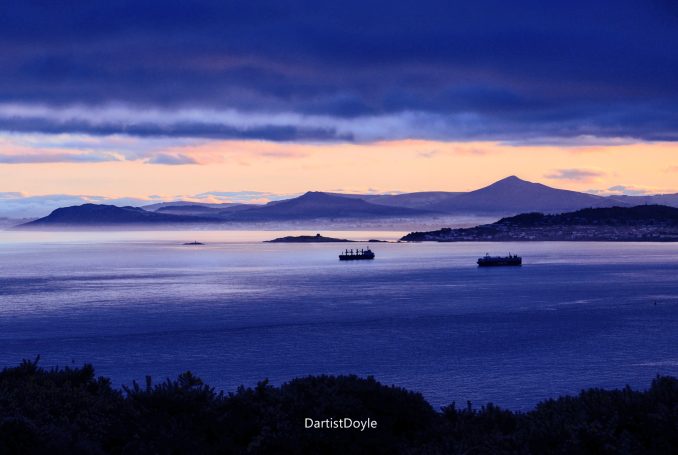 Coucher de soleil sur la mer avec des bateaux et des montagnes à l'horizon.