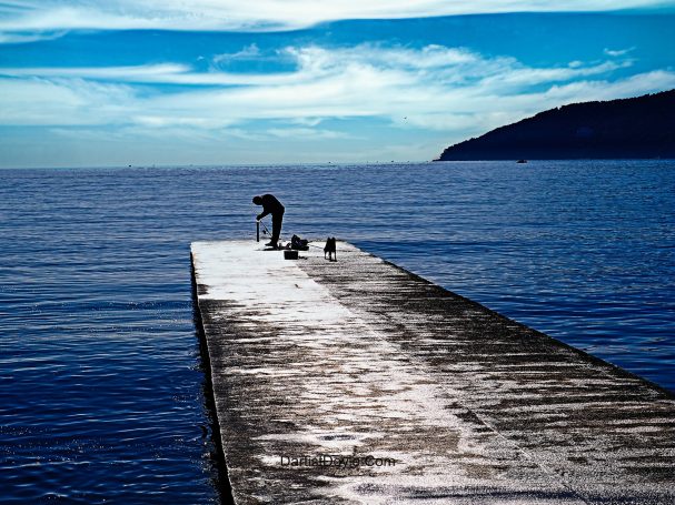 Un homme sur un ponton en bois au bord d'une mer calme, avec des collines à l'horizon.