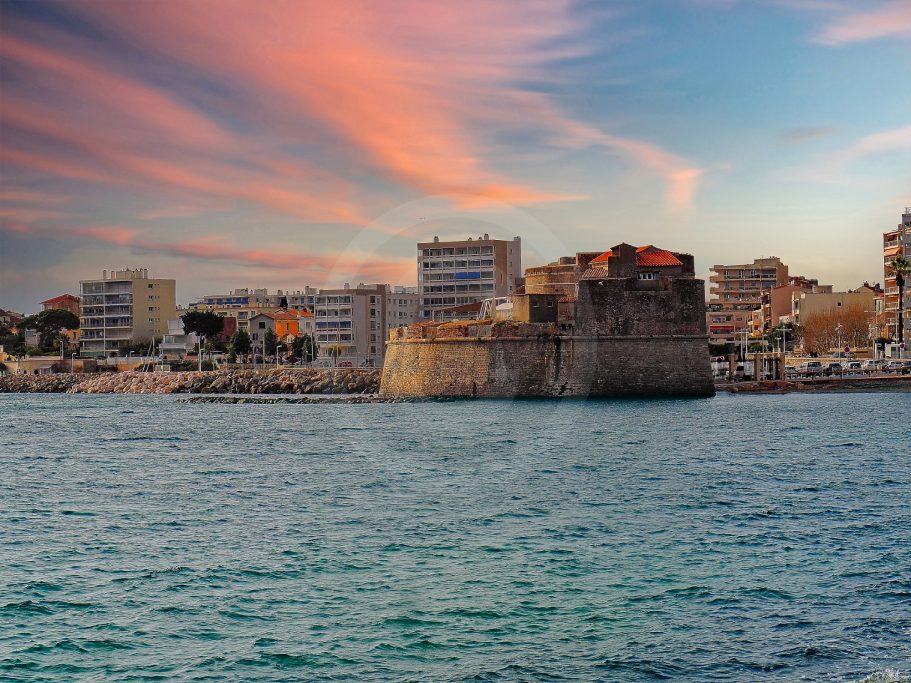 Port avec bâtiments modernes et une forteresse au bord de l'eau sous un ciel coloré.