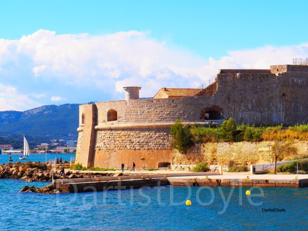 Fortification en bord de mer avec ciel bleu et voilier au loin.