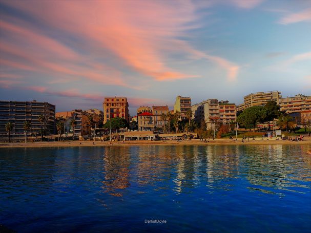Vue d'une plage au coucher du soleil, avec des bâtiments en arrière-plan et un ciel coloré.
