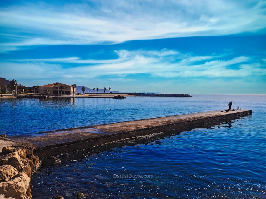 Un homme marche le long d'une jetée au bord d'une eau calme sous un ciel bleu.