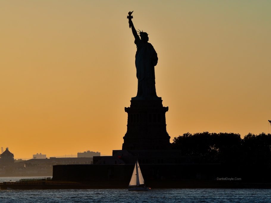Statue de la Liberté au coucher du soleil, silhouette contre le ciel orangé.